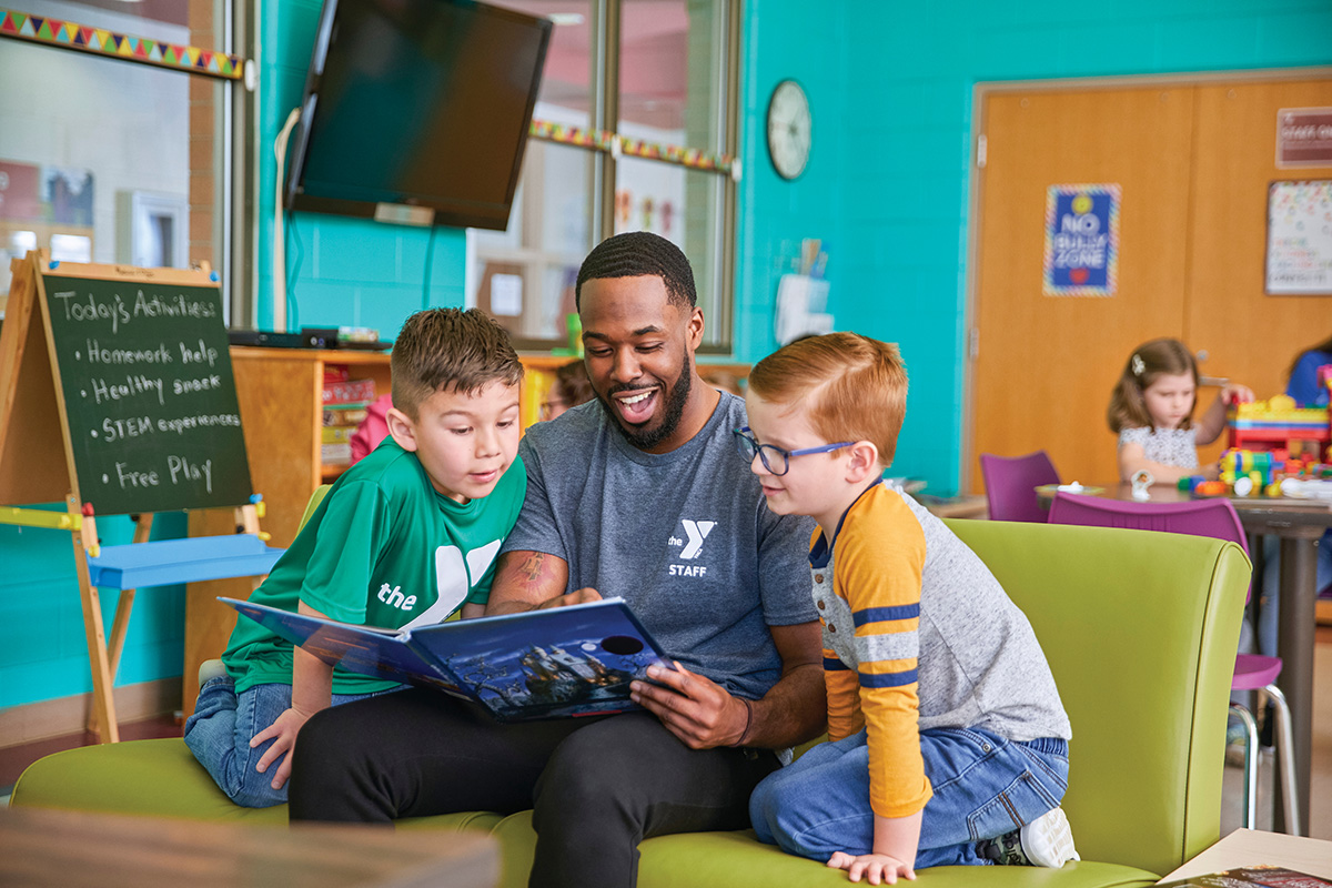 YMCA staff member reading a book to two young boys while seated in a colorful activity room.