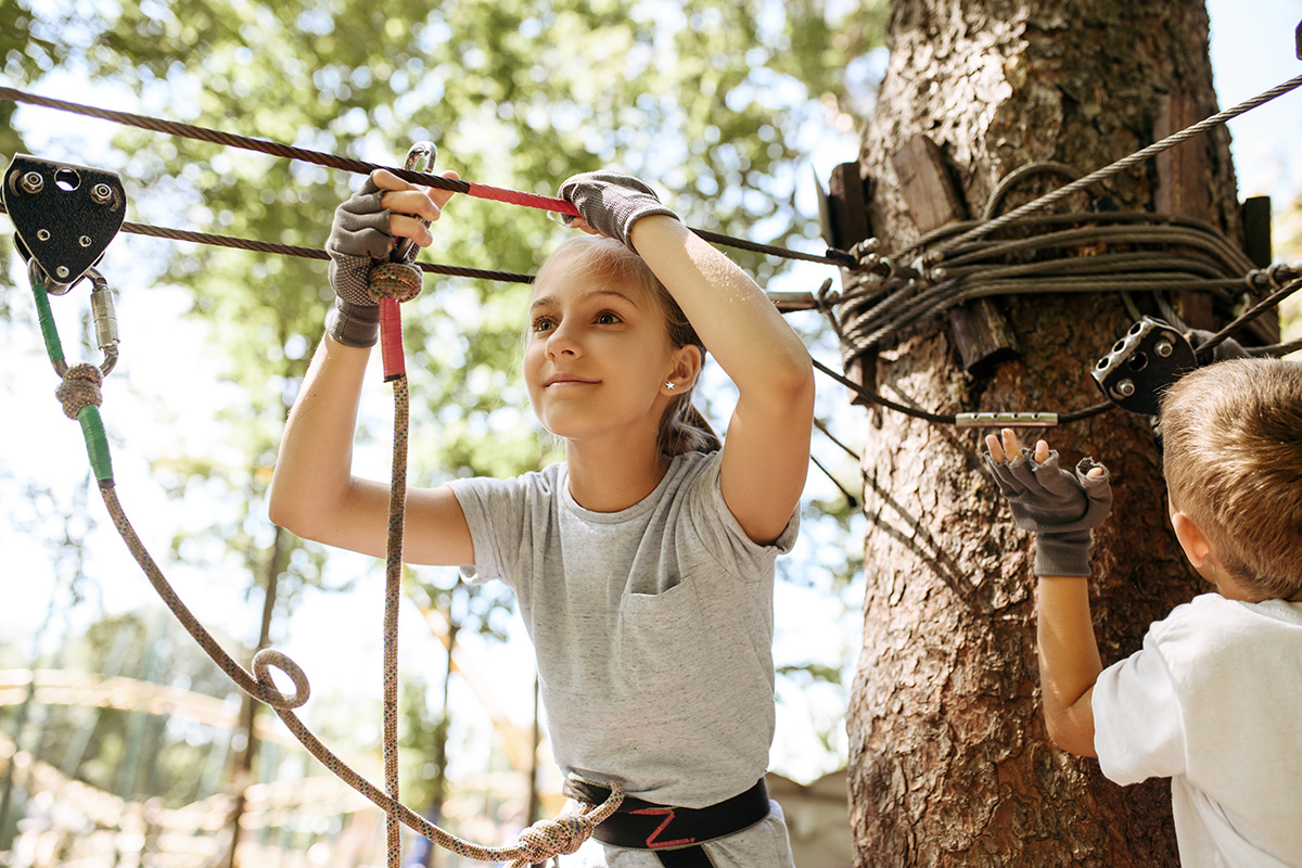 Child wearing gloves and harness navigating a treetop ropes course, surrounded by trees on a sunny day.