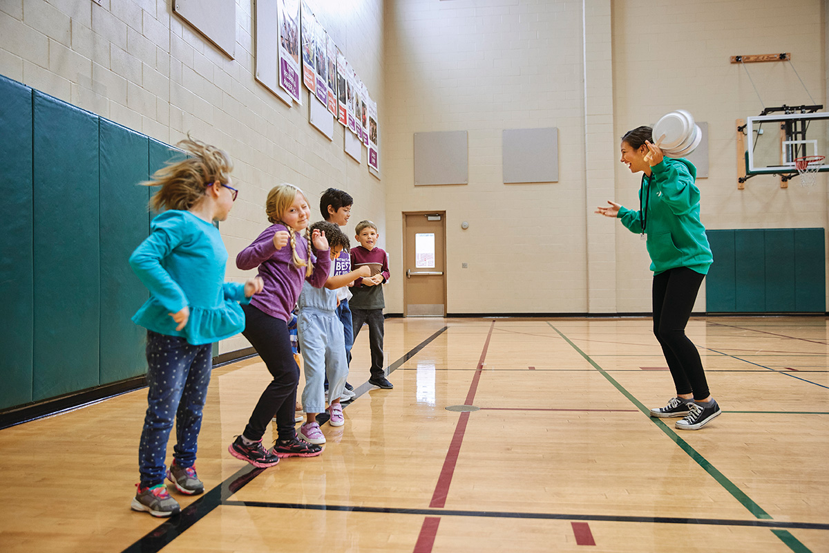 A group of children participates in a gym activity led by an instructor holding plastic discs.