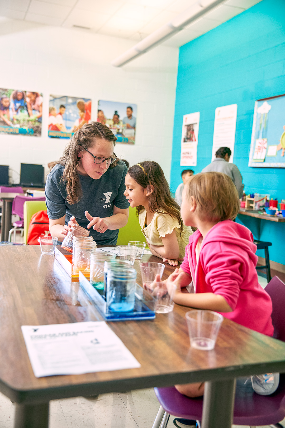 A staff member guides two young girls through a hands-on science experiment with colorful liquids in a classroom.