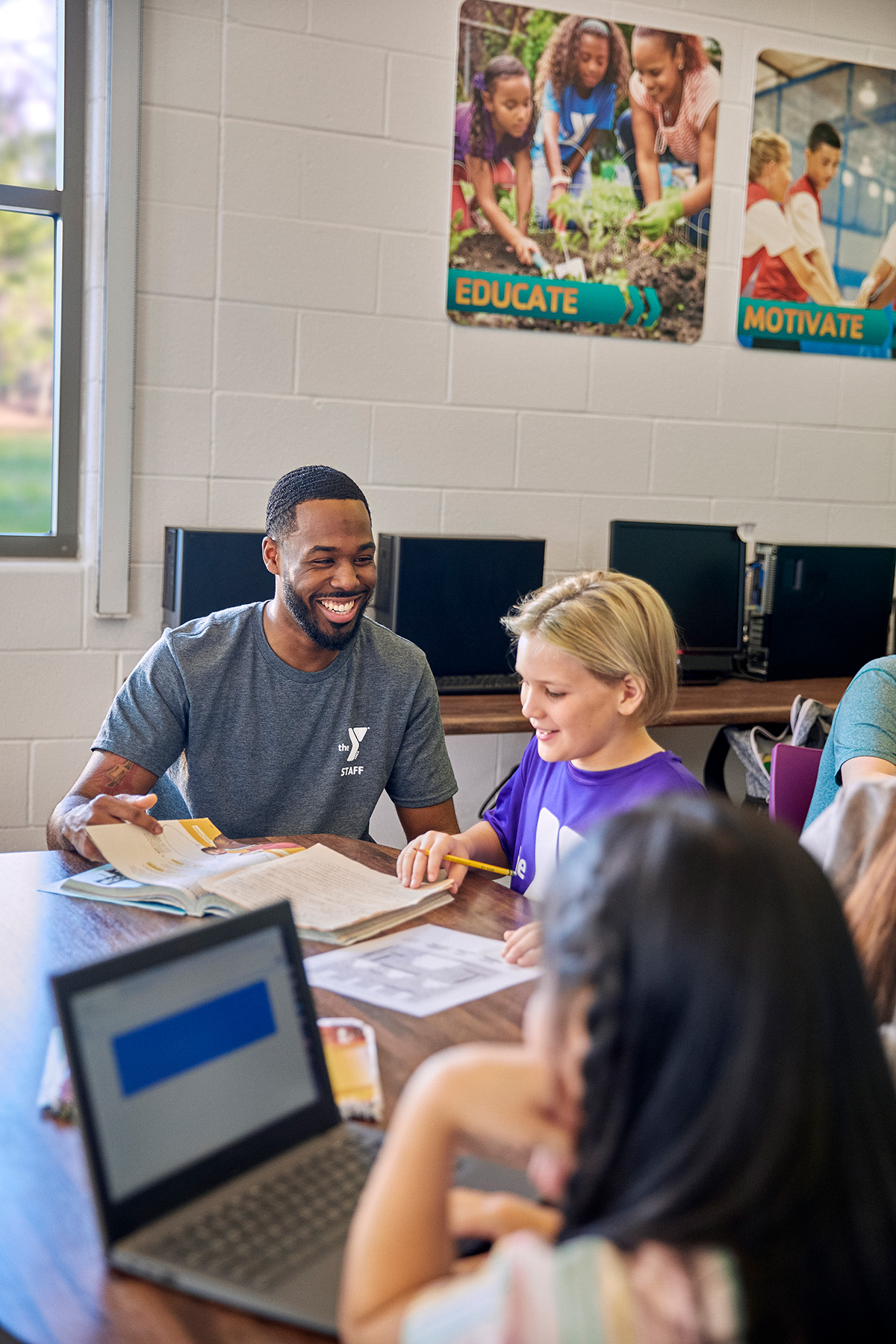 A staff member helps children with homework at a table in a computer lab, smiling and engaging with a student.