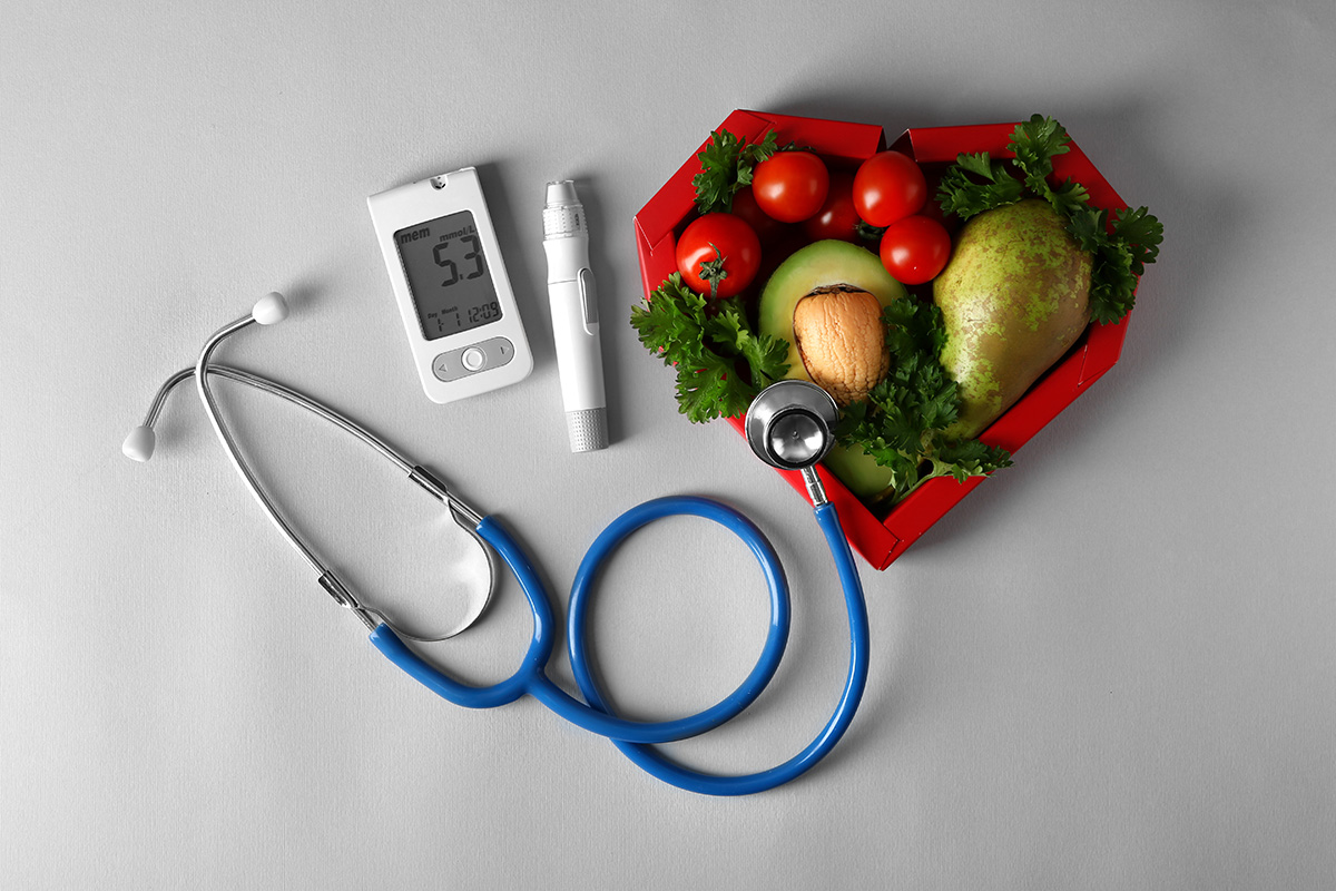 A stethoscope, glucometer, and healthy foods like tomatoes, avocado, and pear arranged in a heart-shaped box, symbolizing health and wellness.