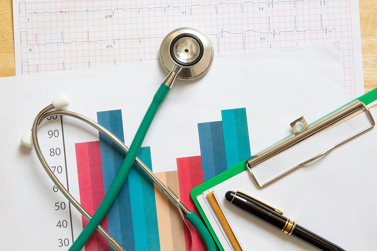 A green stethoscope placed on a table with colorful bar graphs, an ECG chart, and a clipboard with a black and gold pen.