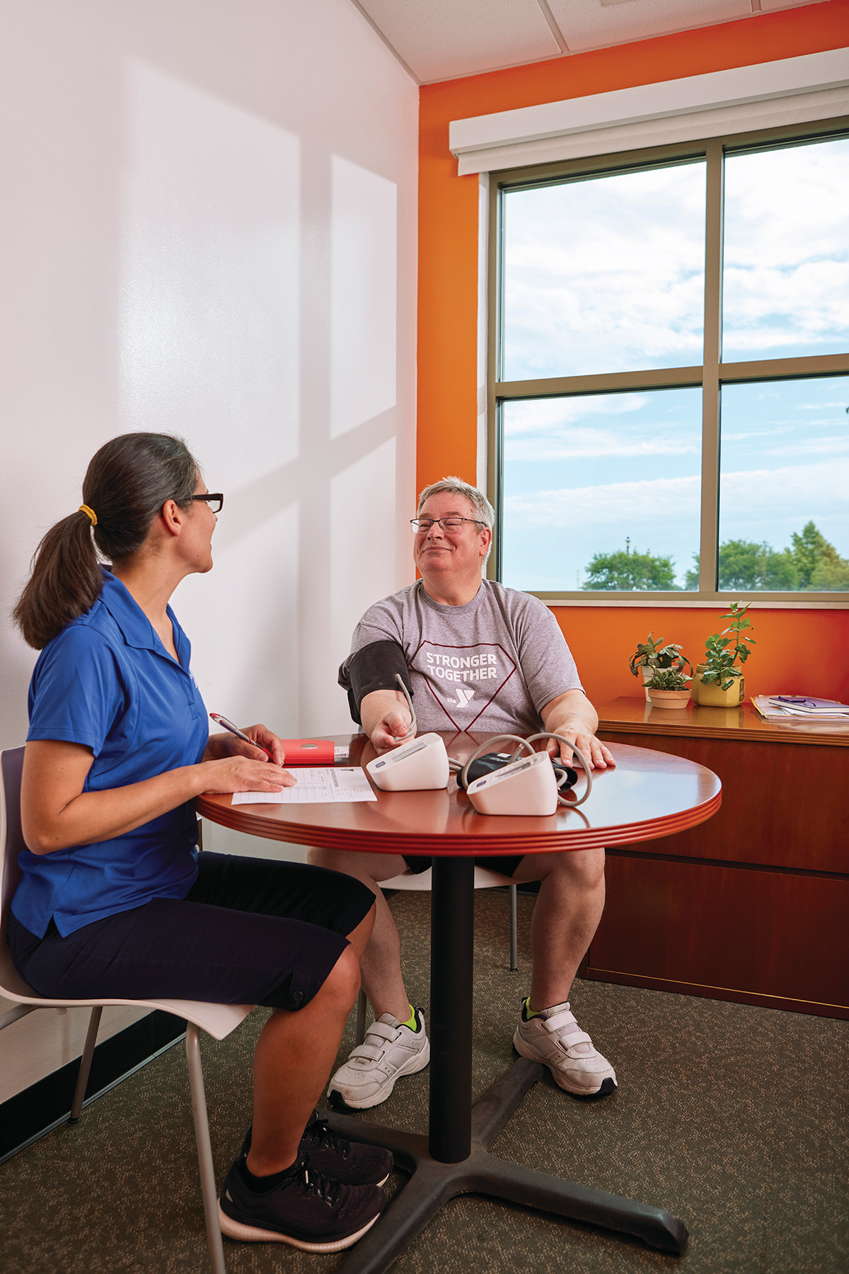 A woman in a blue polo shirt takes notes while measuring the blood pressure of a smiling man sitting at a table in a bright room with an orange accent wall and a window overlooking trees.