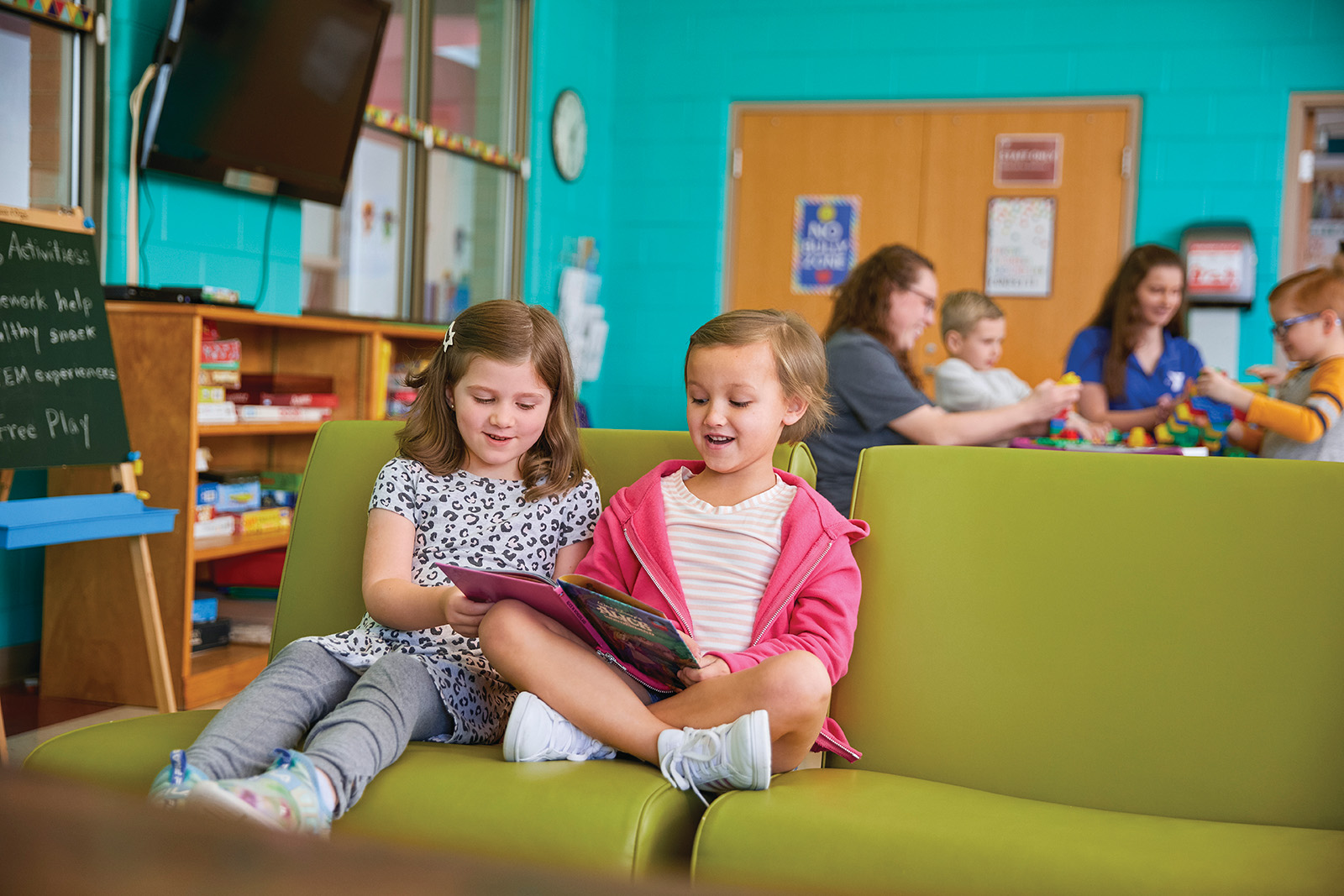 Two young girls read a book together on a green couch in a colorful activity room, while other children play in the background.