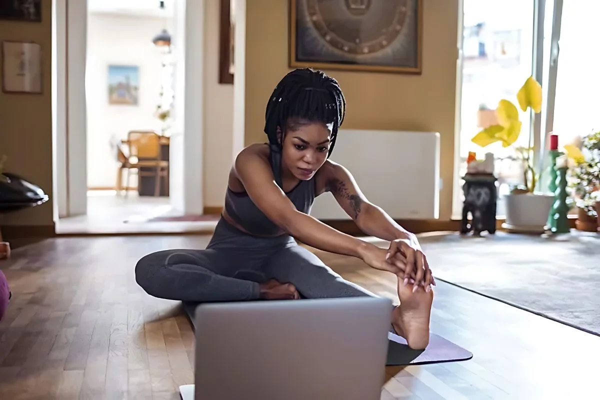 A woman practicing yoga at home, stretching her leg while sitting on a mat and following instructions on a laptop screen. The room is warmly lit, with decor and plants visible in the background.