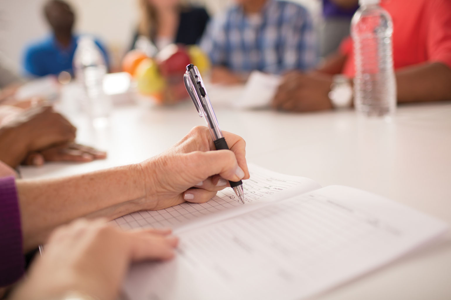 A close-up of a person writing notes in a notebook during a group meeting, with blurred participants and fruit in the background.