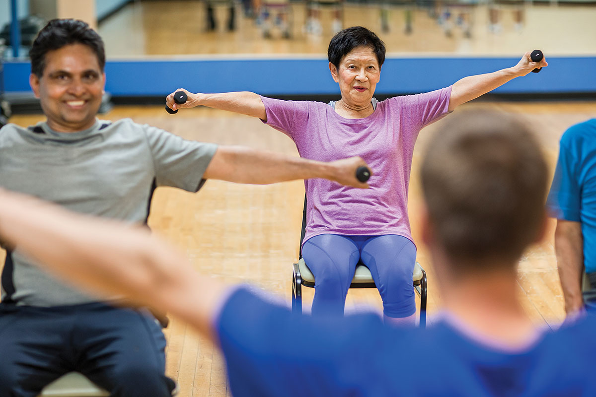 Older adults participating in a seated fitness class, using small dumbbells for arm exercises in a gym with mirrored walls.