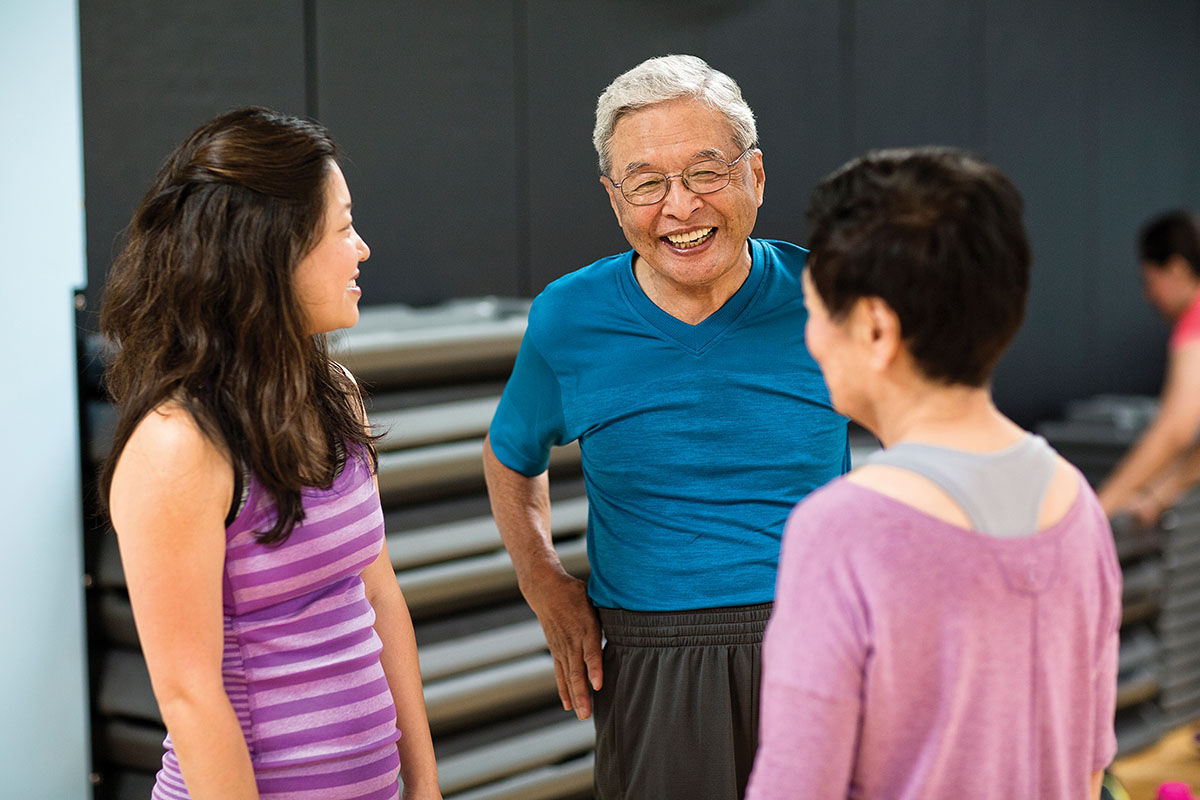 Three adults smiling and chatting in a fitness studio, with stacked exercise mats visible in the background.