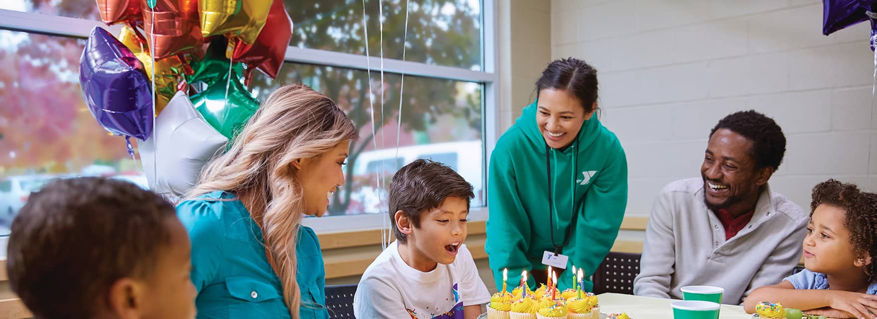 Child blowing out candles on cupcakes during a birthday celebration, surrounded by smiling adults and children, with balloons in the background.