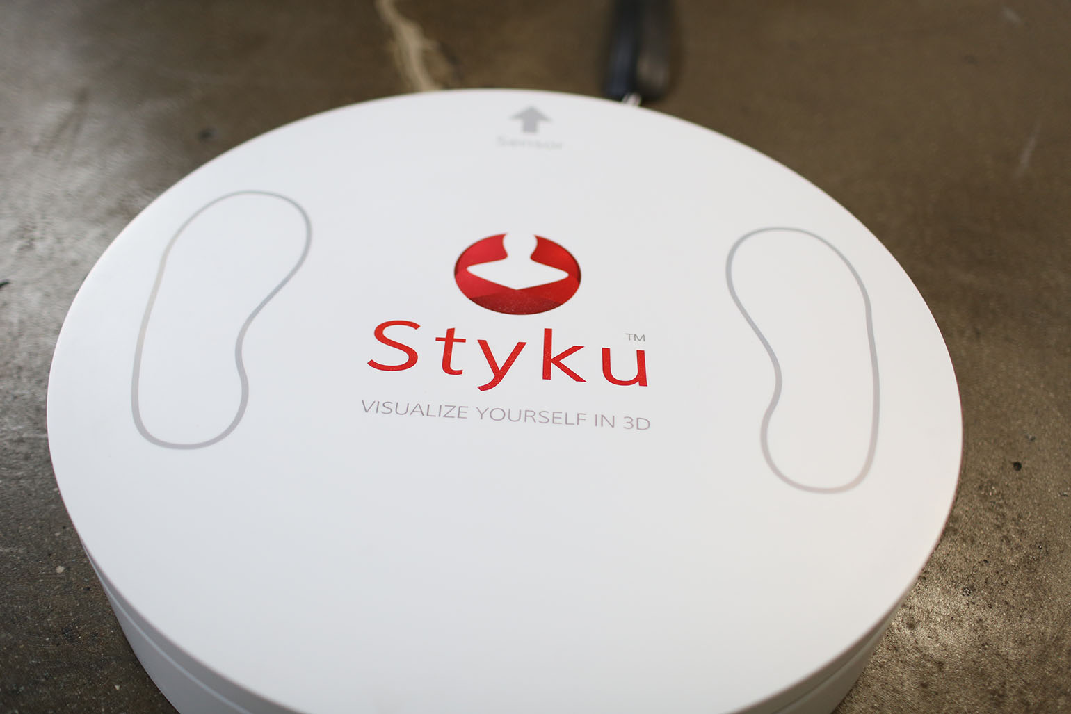 Close-up of a Styku 3D body scanner platform, showing its logo and foot placement outlines on a smooth white surface.