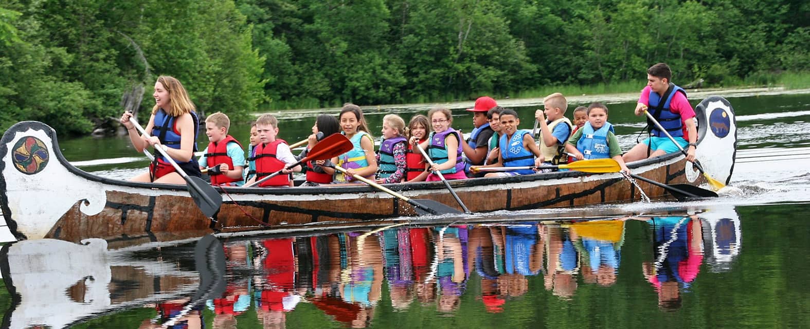 A group of children and adults paddling a large wooden canoe across a calm lake surrounded by lush green trees. The participants wear colorful life vests and are engaged in teamwork as they navigate the water.