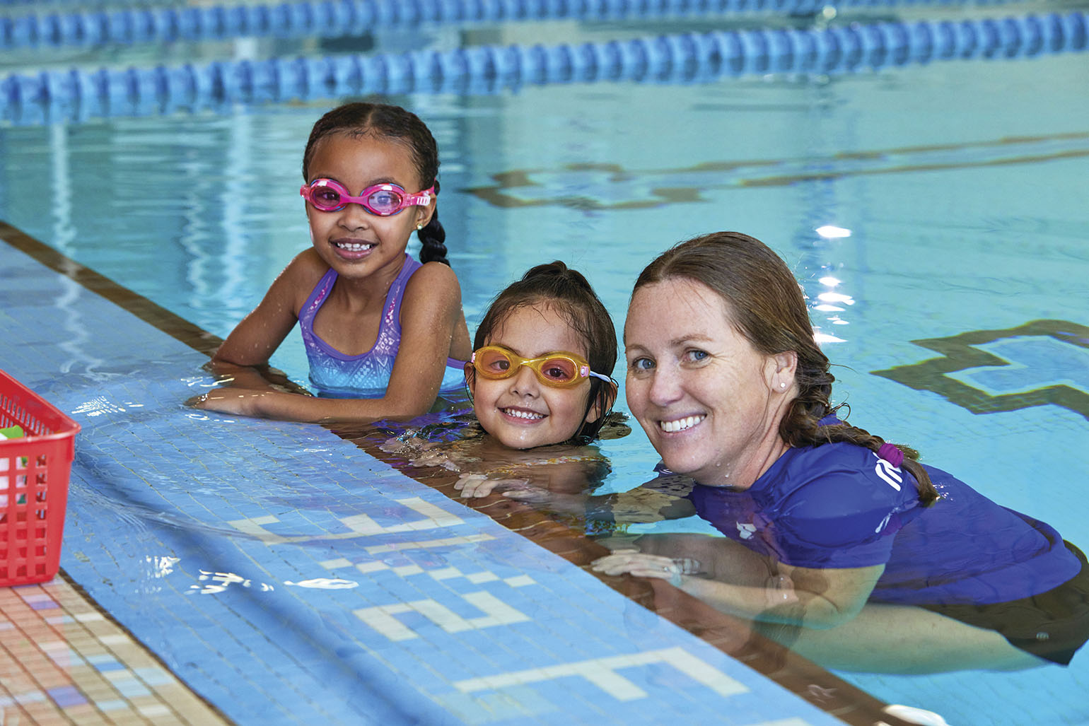 A smiling YMCA instructor teaches two smiling children during their swim lesson at the indoor pool.