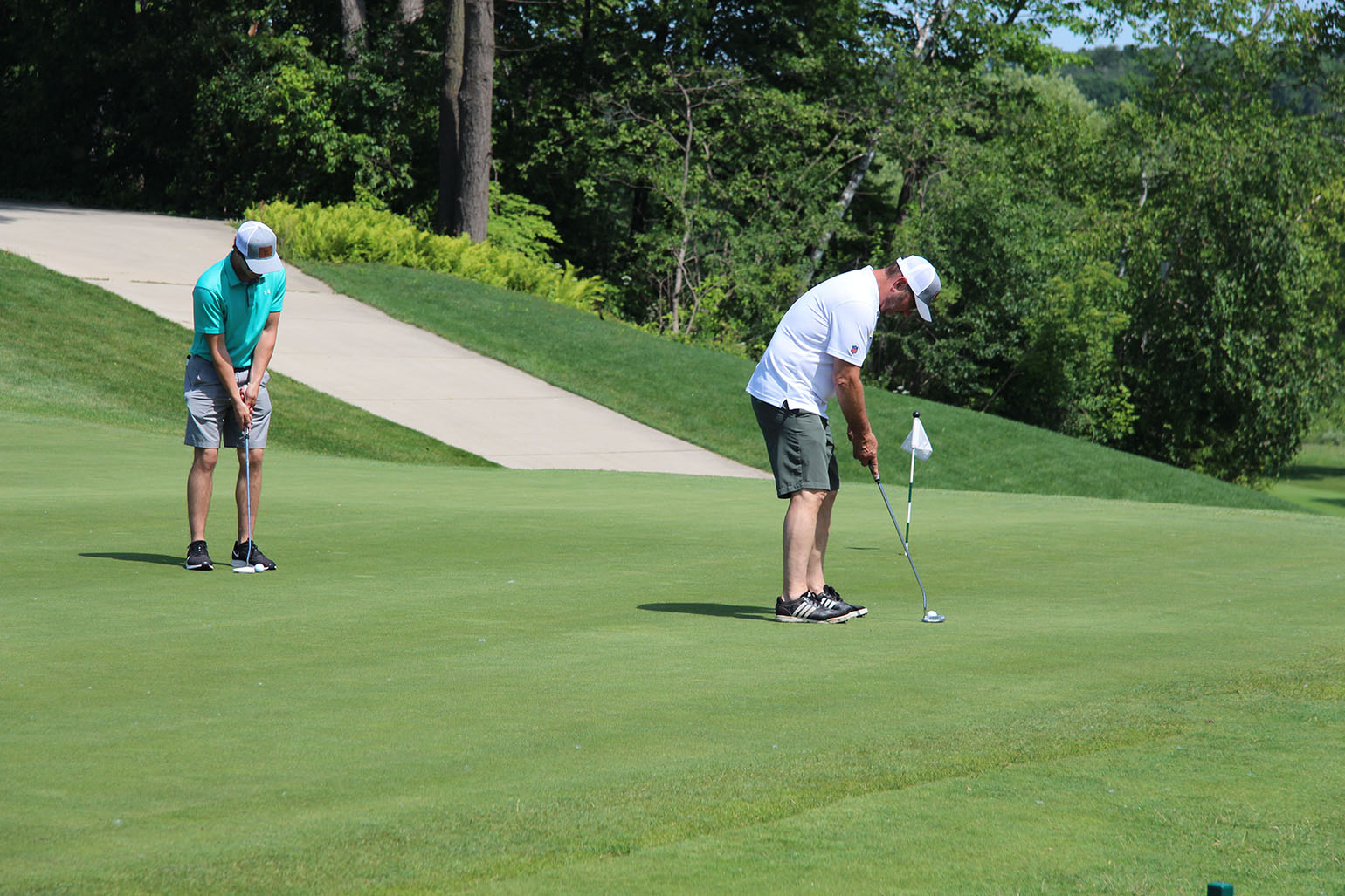 Two men golfing on a sunny day, one lining up a putt while the other prepares to play on a well-maintained green surrounded by trees.