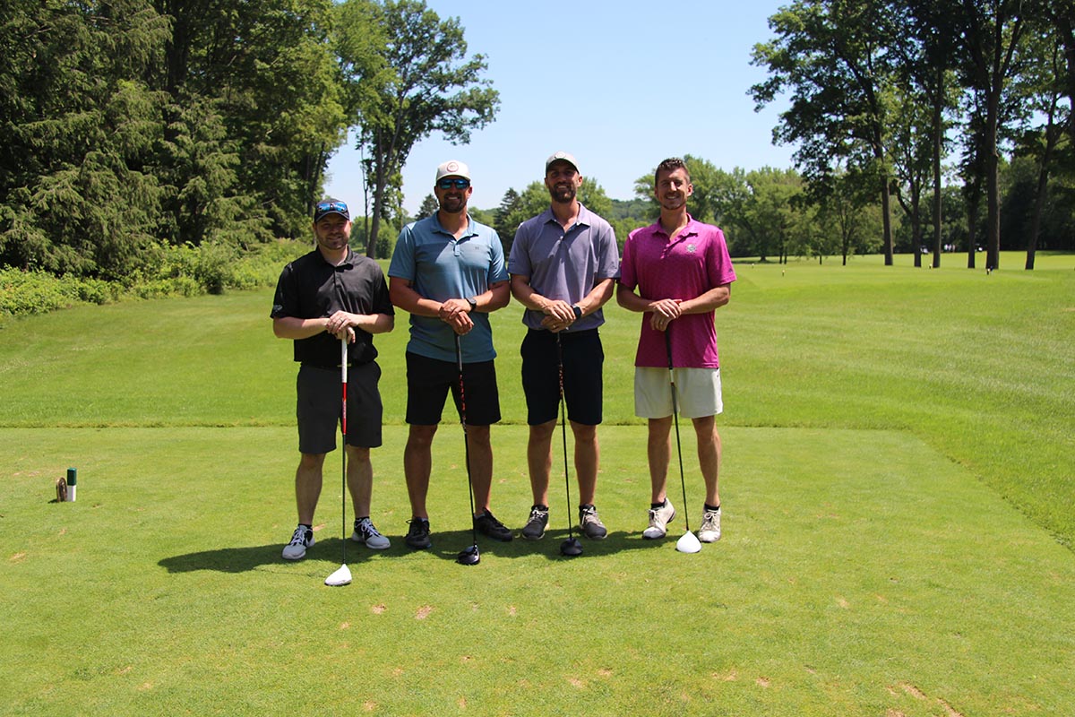 Four men standing together on a golf course, holding clubs and smiling, with a scenic green backdrop and trees in the distance.