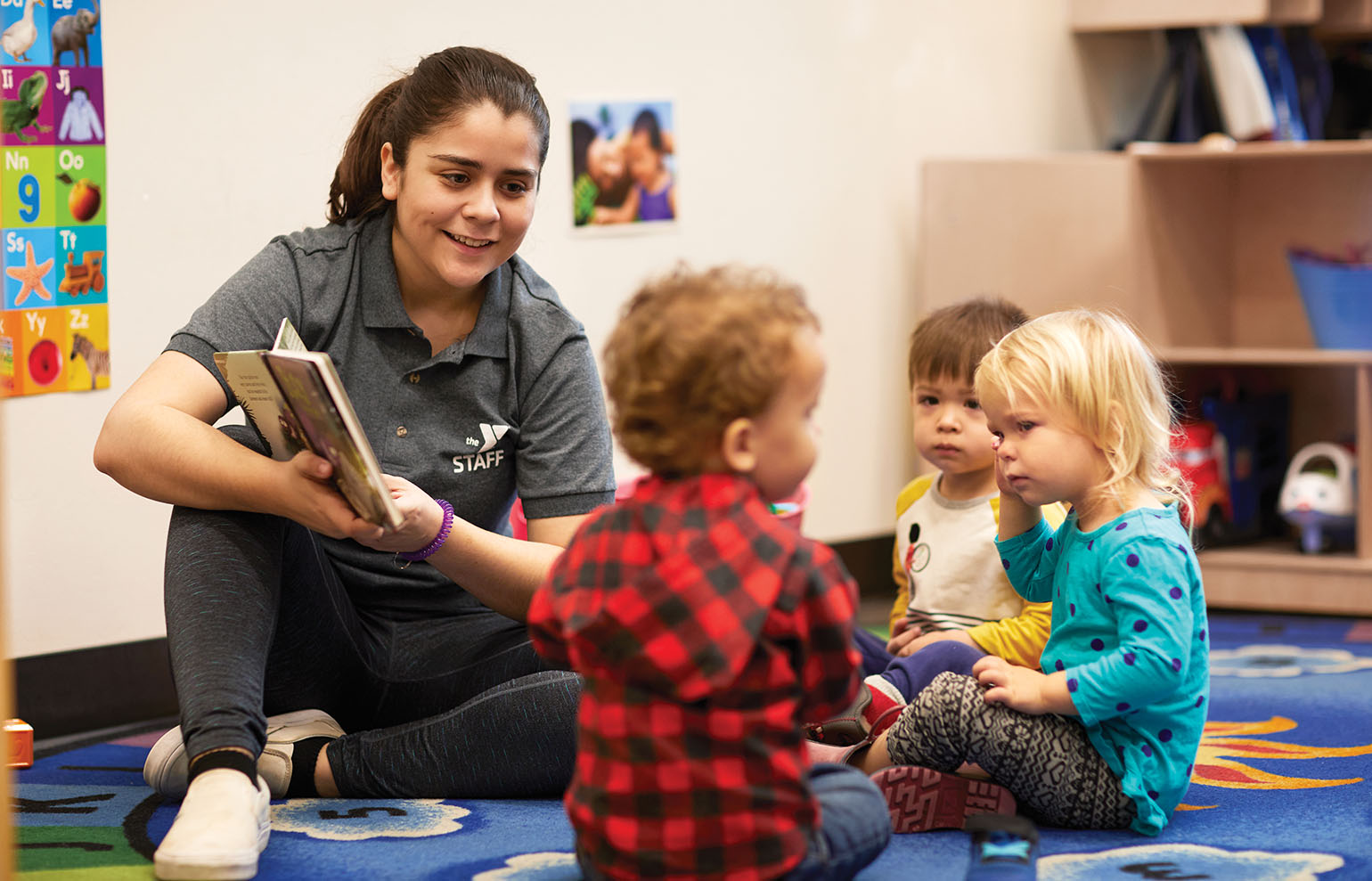 A childcare staff member reads a book to a group of toddlers sitting on a colorful classroom rug.