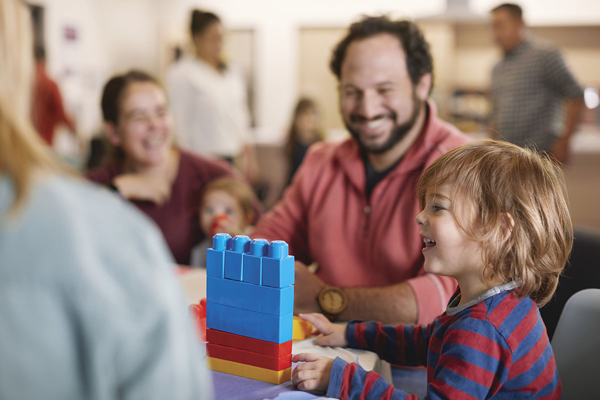 A smiling child builds with colorful blocks at a table while adults interact in a family-friendly setting.