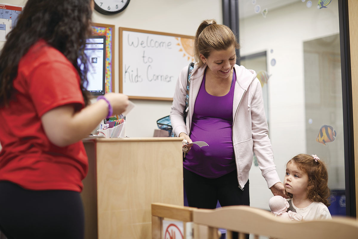 A pregnant mother checks in at a childcare center with her young daughter holding a doll.