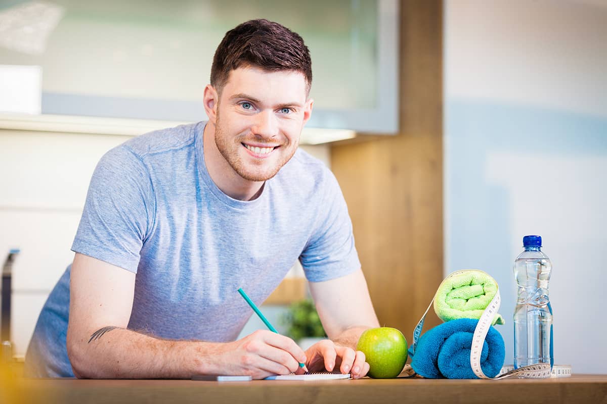 Smiling man writing in a notebook at a kitchen counter with a green apple, water bottle, rolled towels, and measuring tape beside him.