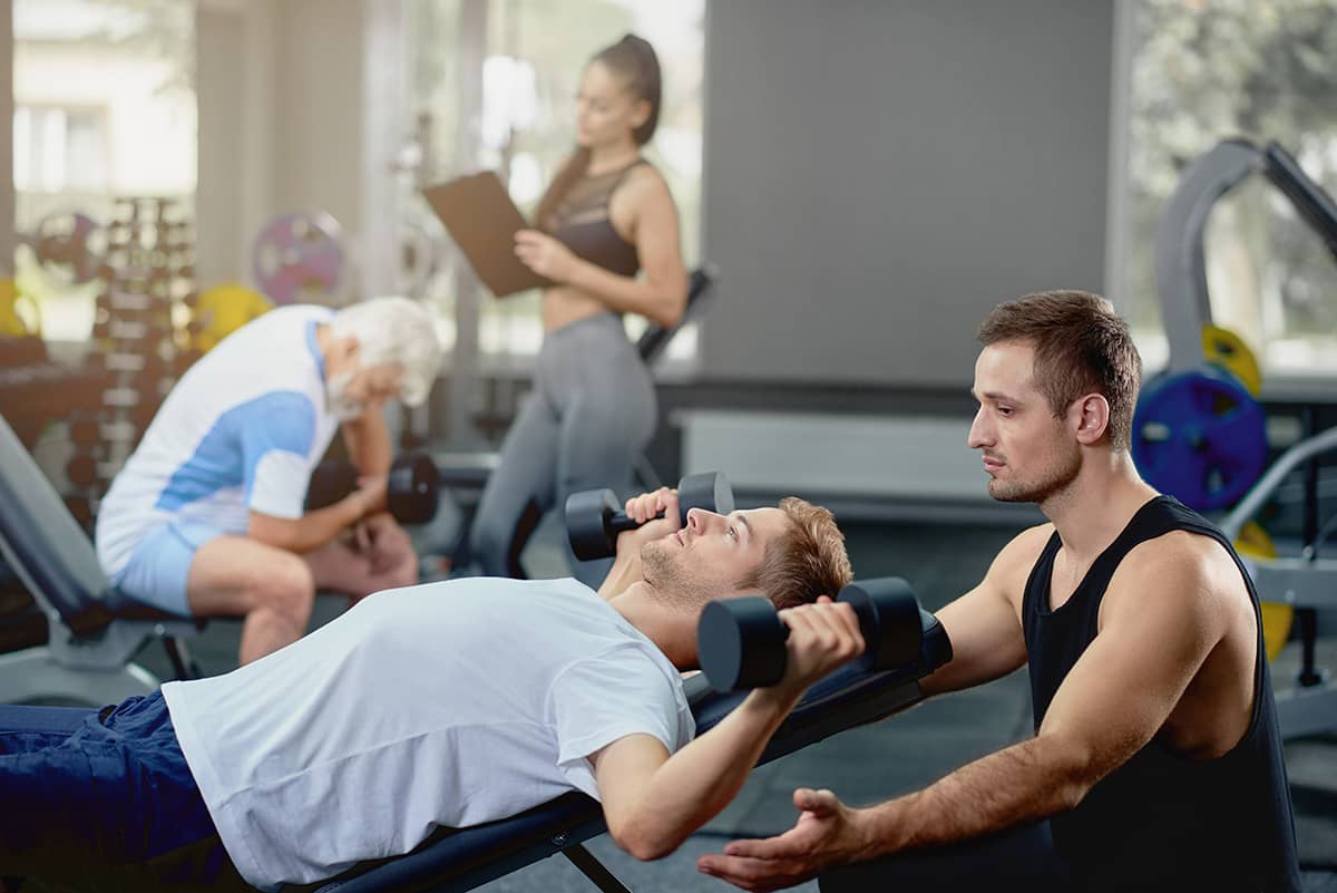 A man lying on a bench lifting dumbbells while being guided by a trainer in a gym. In the background, other individuals exercise, including an older man and a woman holding a clipboard.