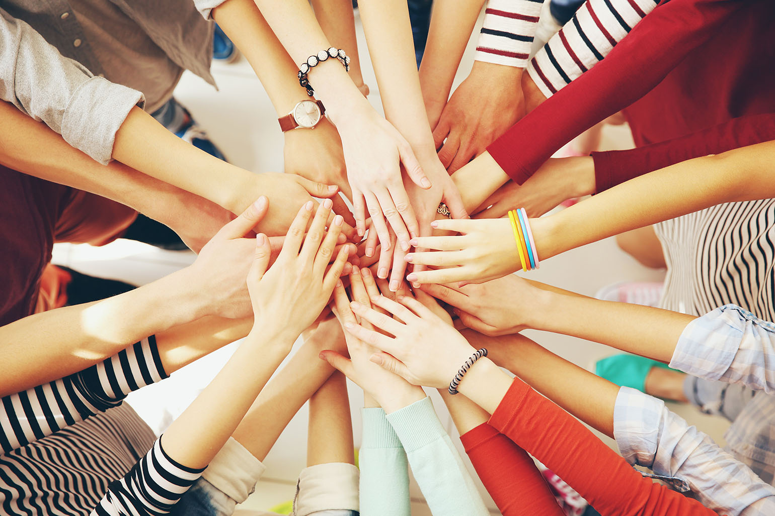 A group of diverse hands stacked together in the center, symbolizing teamwork and unity. The hands belong to individuals wearing colorful clothing, bracelets, and watches.