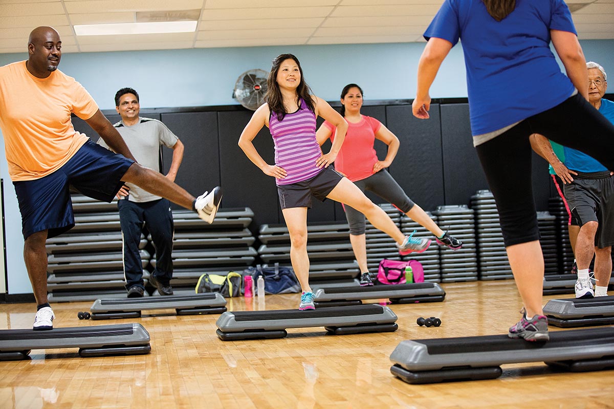 Group fitness class participants performing step aerobics on platforms in a bright exercise studio.