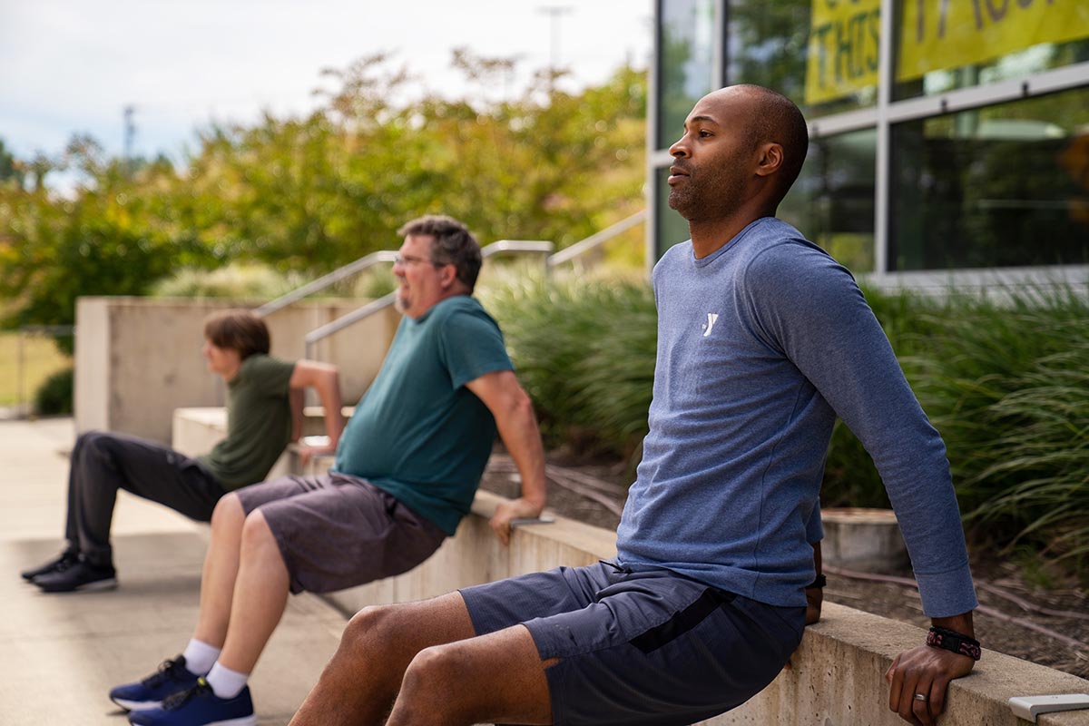 Three men of different ages perform tricep dips on outdoor steps, exercising together in a sunny, grassy setting.