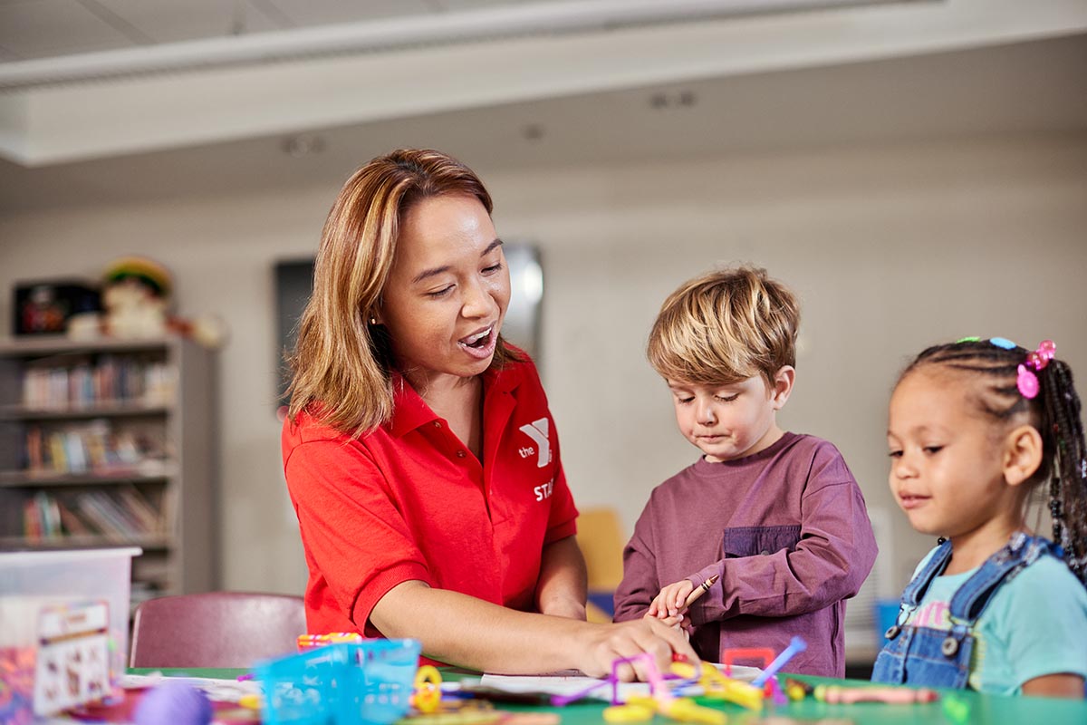 YMCA staff member in a red shirt guiding two young children in an arts and crafts activity at a table in a classroom setting.