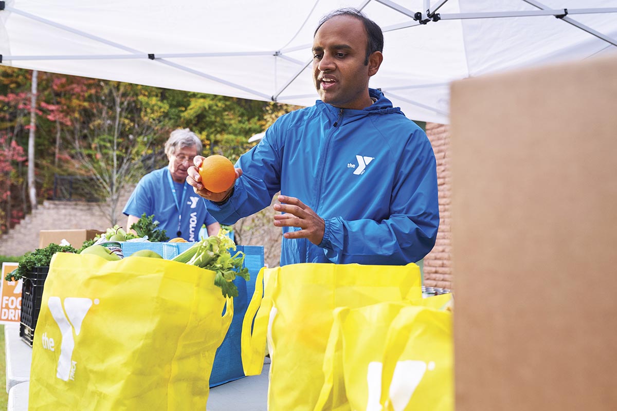 A volunteer in a blue jacket holds an orange while organizing fresh produce in yellow bags during an outdoor food drive.