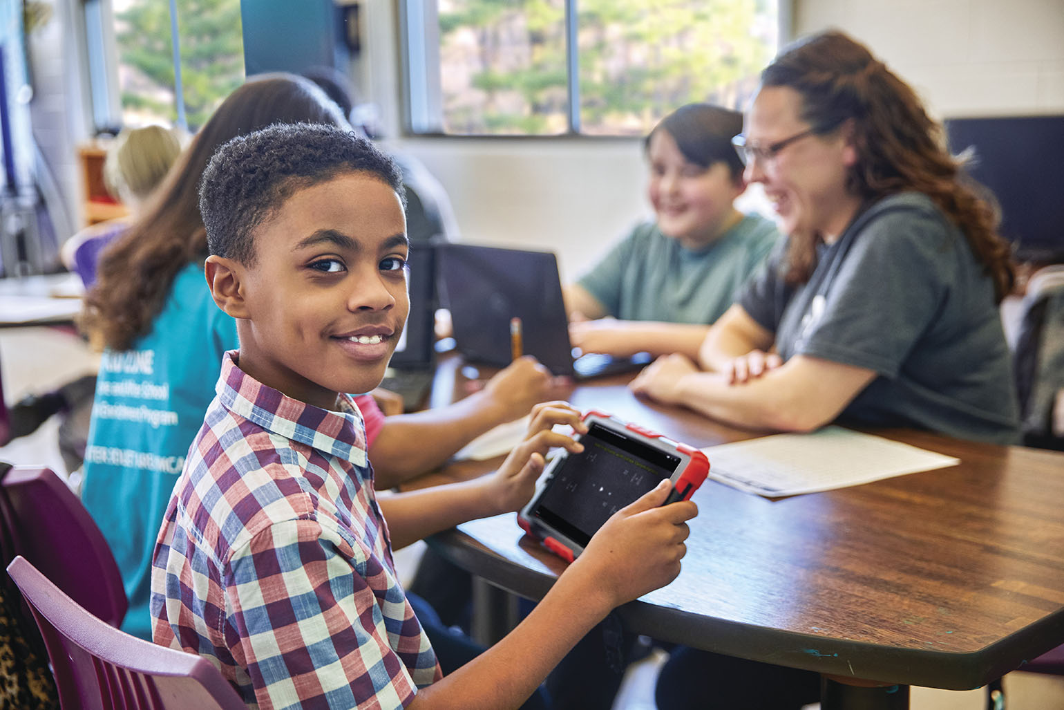 A smiling child looks at the camera while holding a tablet as Y staff workers help other children with their homework.