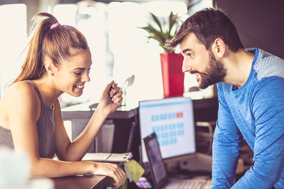 A woman and a man engage in a friendly conversation at a desk with a laptop and calendar on the screen, in a bright indoor space.