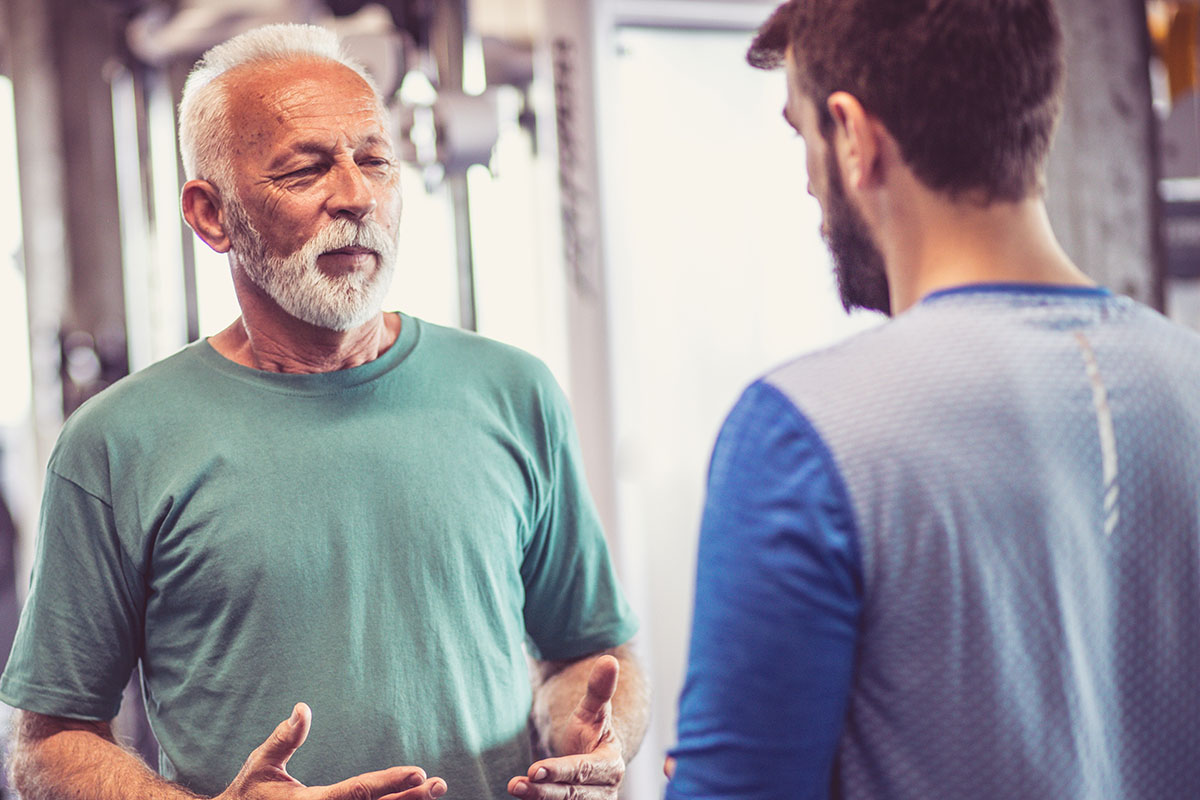 Senior man talking to a younger man in a gym surrounded by exercise equipment.