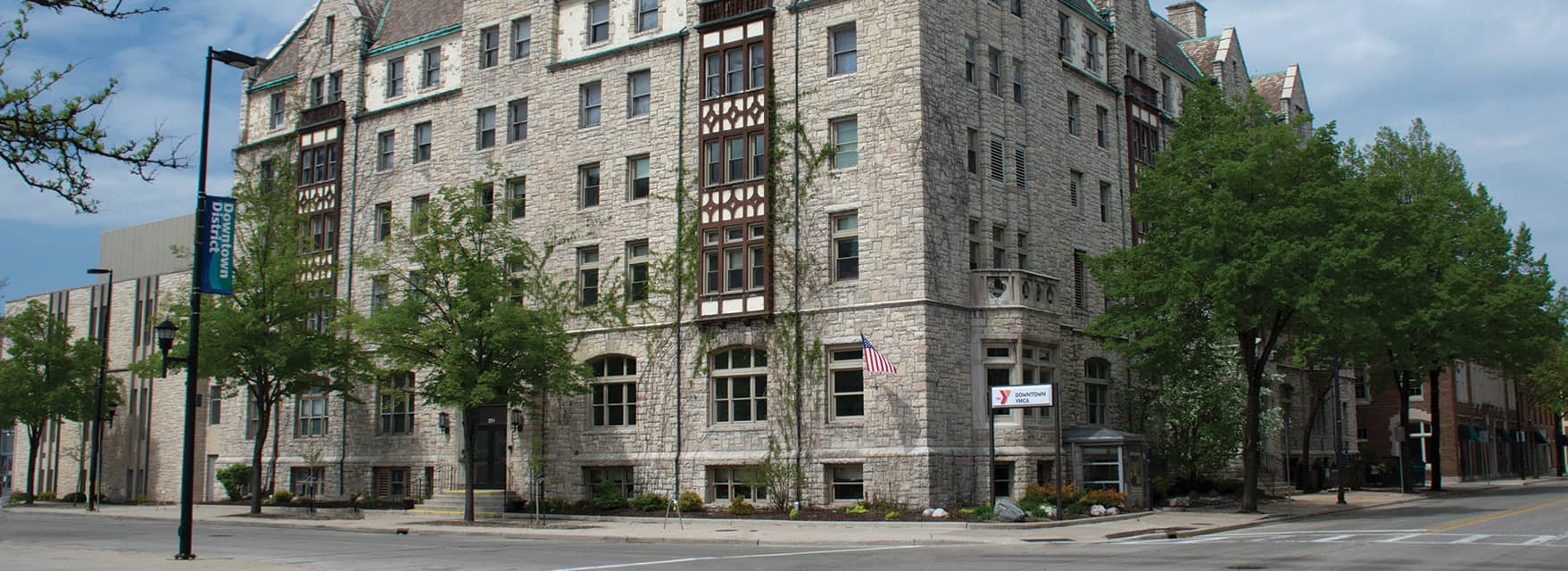 Historic stone YMCA building on a corner street, surrounded by trees and a sign reading "Downtown YMCA" near the entrance.