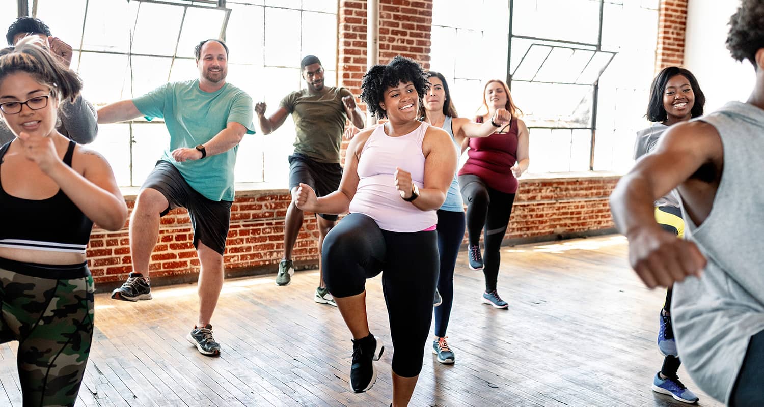 A diverse group of adults participating in a high-energy fitness dance class in a studio with brick walls and large windows. Participants are smiling and lifting their knees during a choreographed routine. The room is bright with natural light streaming in.