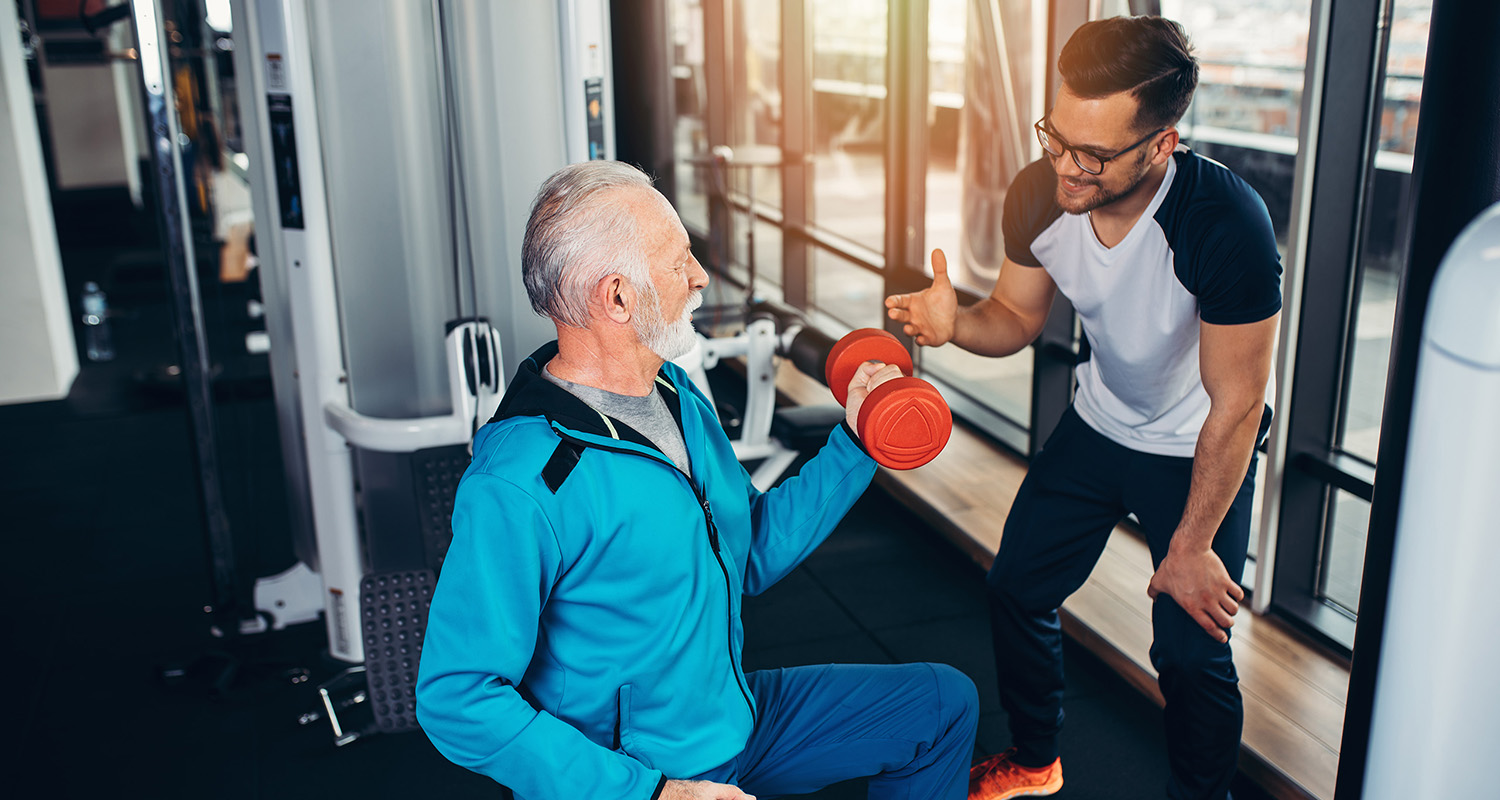 An older man lifting a dumbbell while receiving encouragement from a trainer in a modern gym setting.