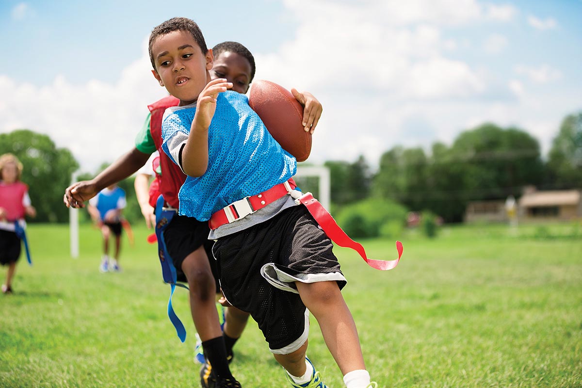 Children play flag football on a sunny day, with one child running while holding a football as others chase behind.