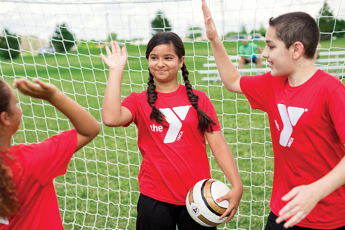 Young soccer players wearing red YMCA shirts celebrate with high-fives near a goal net on a grassy field.