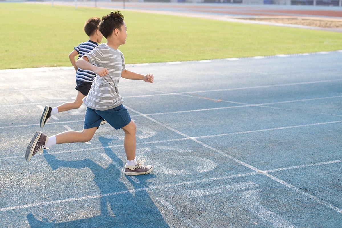 Two children run on an outdoor blue track near a grassy field under bright sunlight.