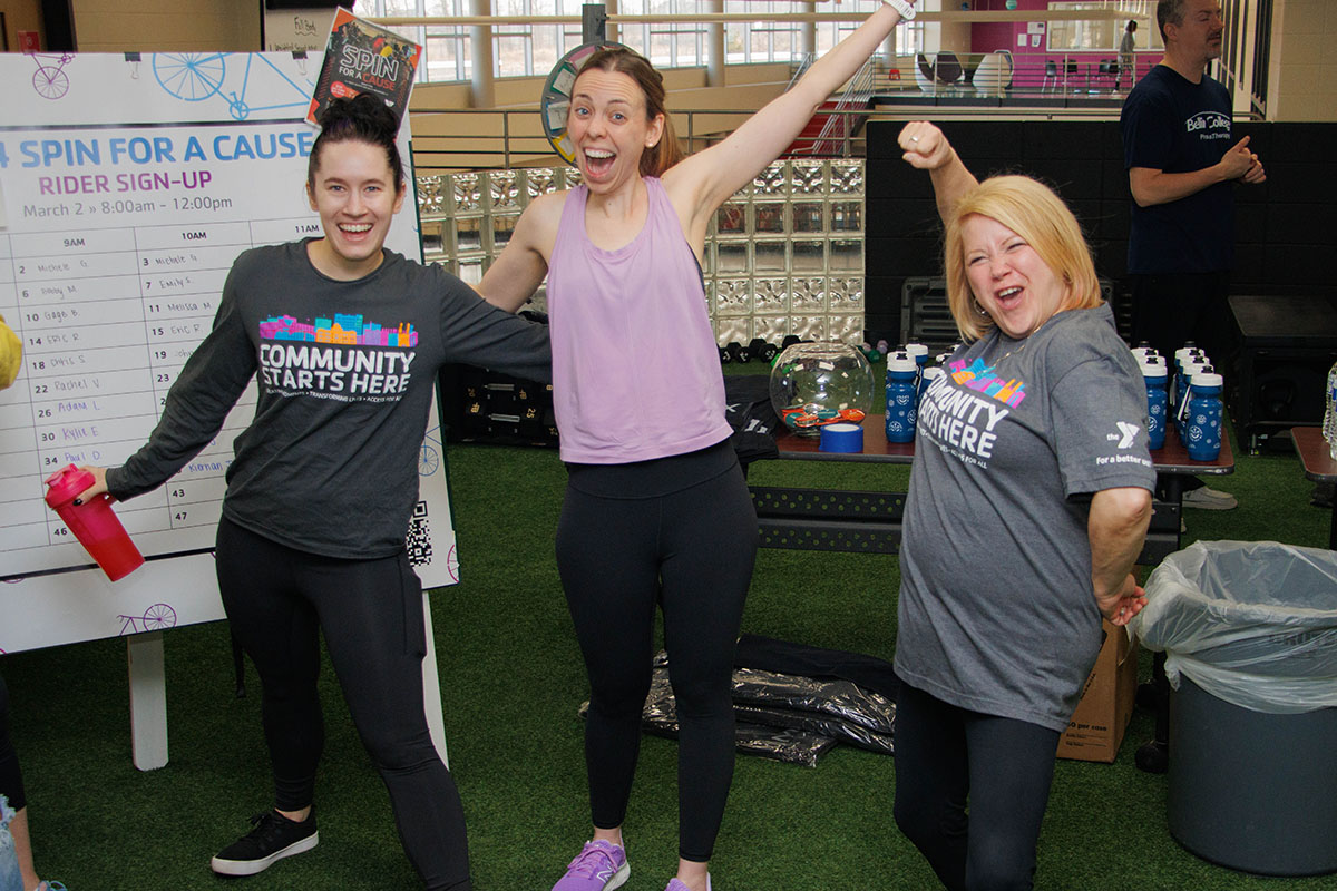 Three enthusiastic women posing and smiling at a YMCA spin event, with a "Spin for a Cause" sign and gym equipment in the background