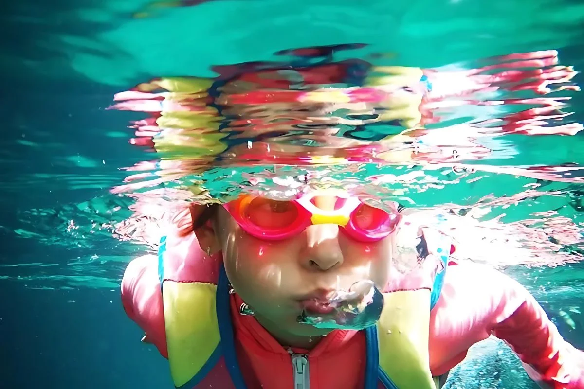 A child in pink goggles and a life jacket blows bubbles underwater in a brightly lit pool.