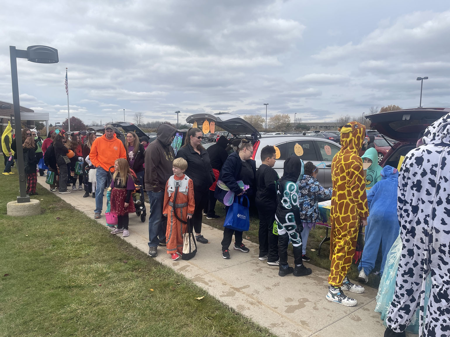 Children and families dressed in costumes participating in a trunk-or-treat event outdoors, with decorated vehicles and overcast skies.