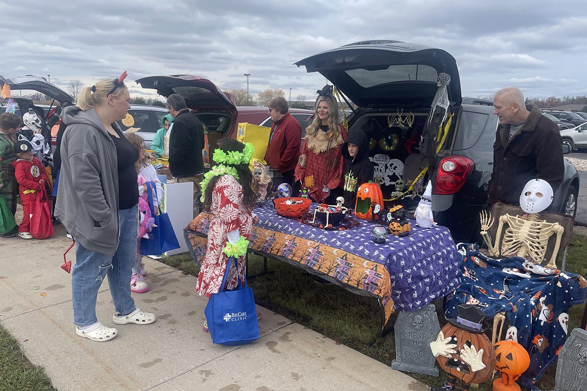 People gathered at a Halloween-themed trunk-or-treat event, with decorated car trunks displaying spooky decorations and children in costumes collecting treats.