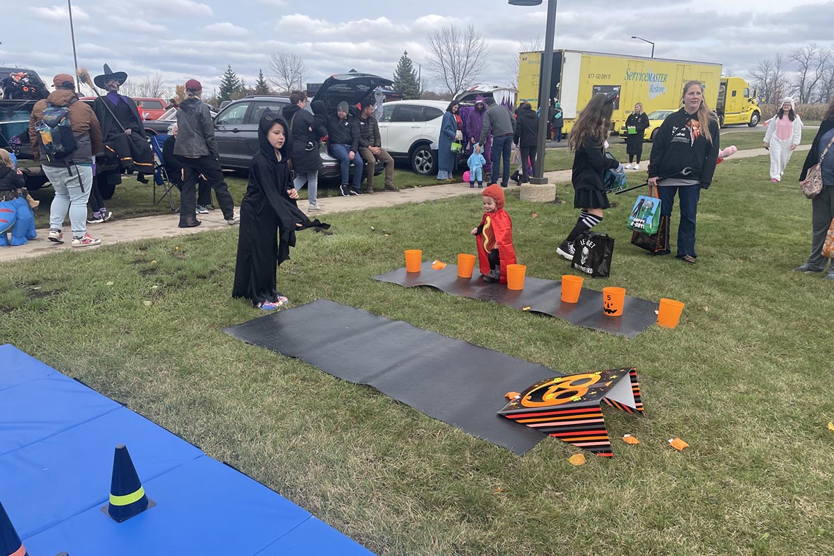 Children dressed in Halloween costumes participate in an outdoor game, tossing items into orange pumpkin buckets on a grassy field, while adults and other costumed children look on during a festive event.