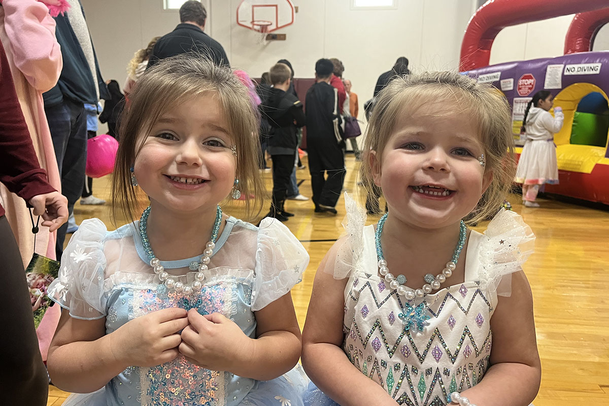 Two young girls smiling in princess costumes with beaded necklaces at an indoor event, surrounded by other attendees and inflatable activities.