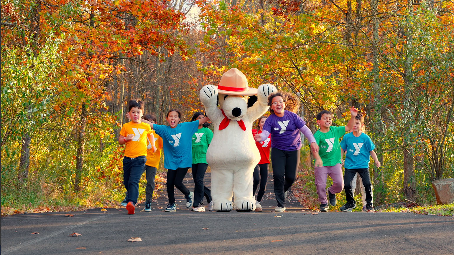 A group of children wearing colorful YMCA shirts joyfully run alongside a large Snoopy mascot dressed as a park ranger on a scenic autumn trail surrounded by vibrant fall foliage.