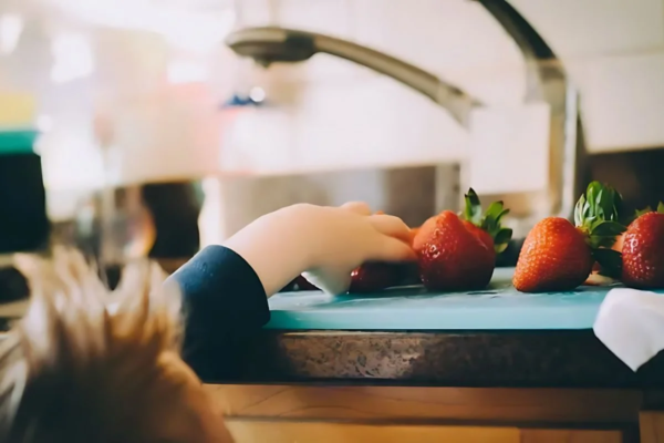 A child’s hand reaches for fresh strawberries placed on a countertop in a brightly lit kitchen.