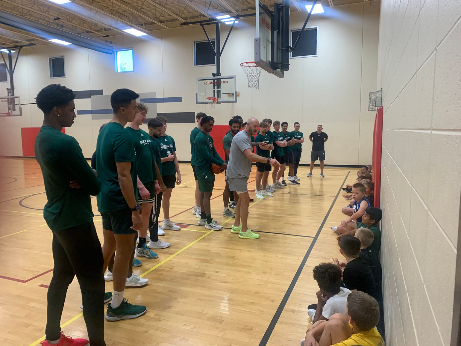 Basketball players in green shirts interacting with a group of children seated against the wall in a gymnasium during a sports clinic.