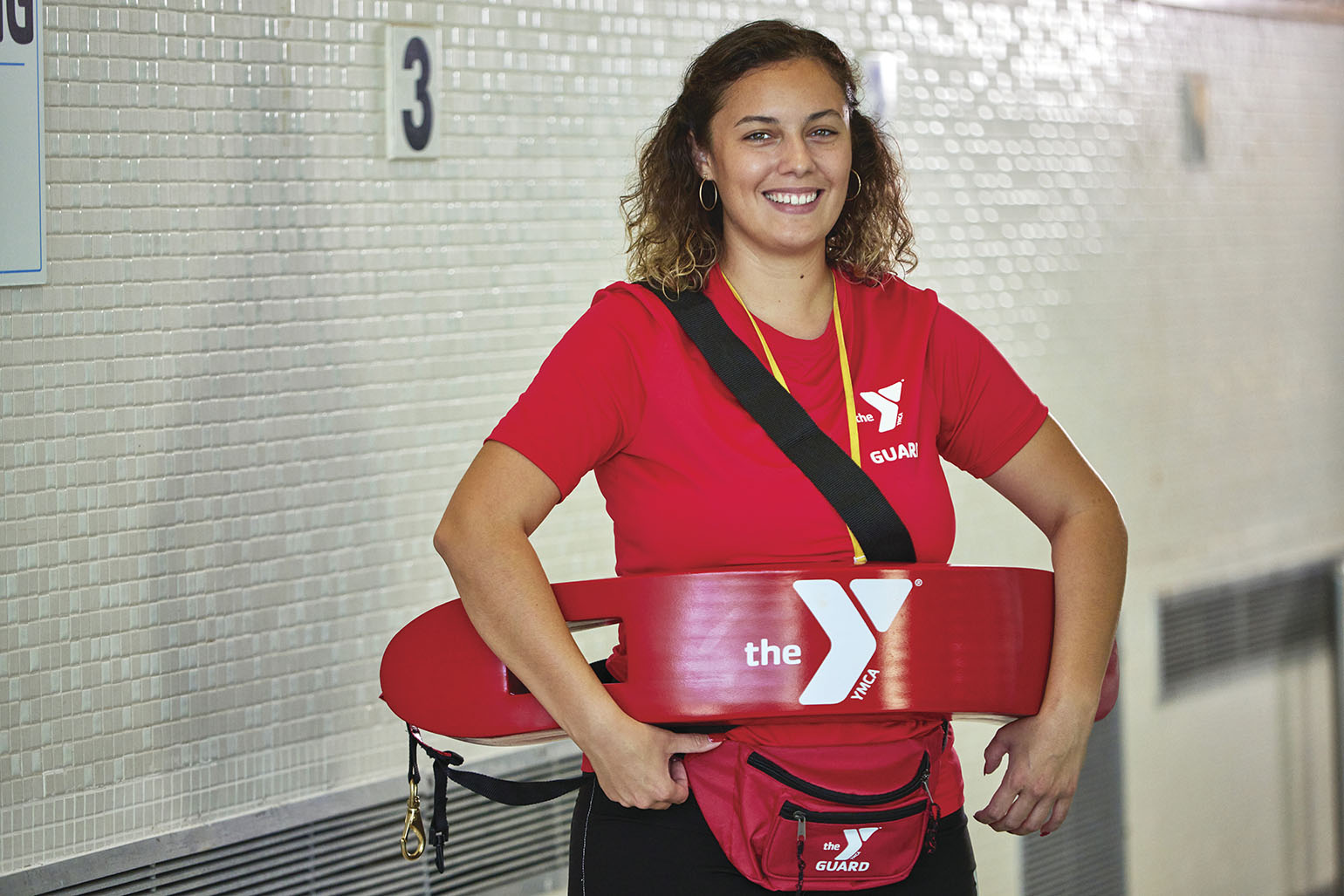 A smiling lifeguard in a red YMCA uniform holding a rescue tube, standing by an indoor swimming pool.