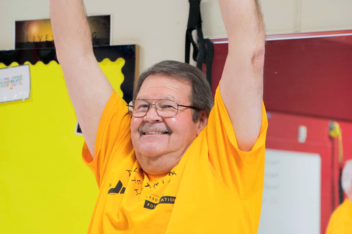 Smiling older man in a bright yellow shirt raising his arms during an indoor fitness activity, expressing energy and joy.