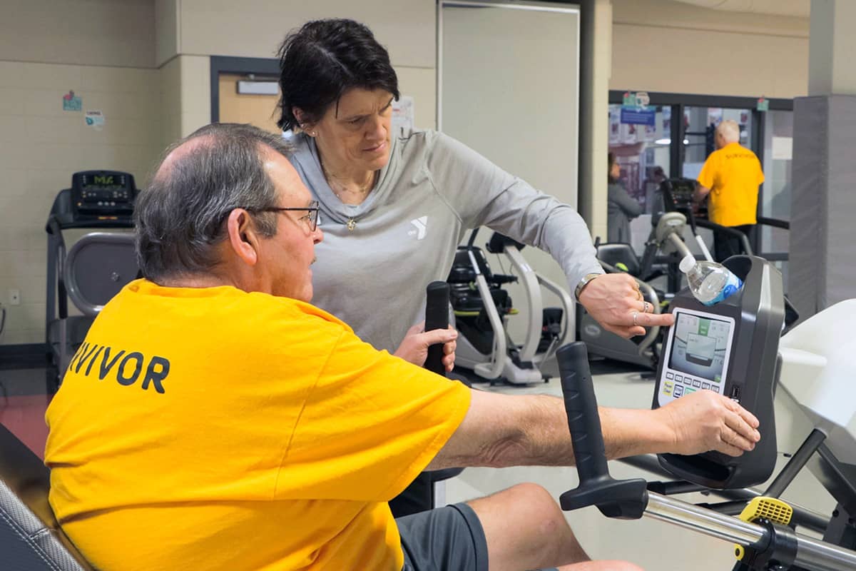 Trainer assisting a man wearing a 'Survivor' shirt with a recumbent exercise bike in a gym setting.